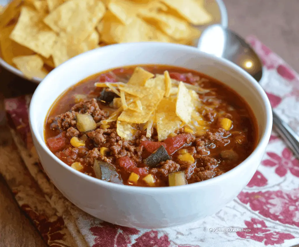 Hearty gluten-free taco soup in a bowl topped with fresh cilantro, shredded cheese, and sliced jalapenos, served with tortilla chips on the side.